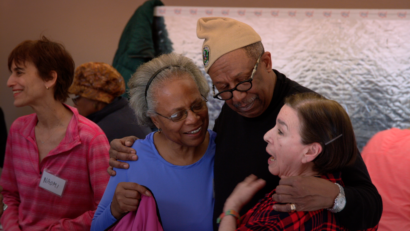 Dark-haired woman dressed in a pink top look to her right smiling. An elderly Black man in a light colored knitted beanie wearing glasses hugs two women. One woman who is Black on his right is gray haired wearing a blue shirt, smiling. The other elderly woman is white with long dark hair speaking. The man's arms encircle both women.